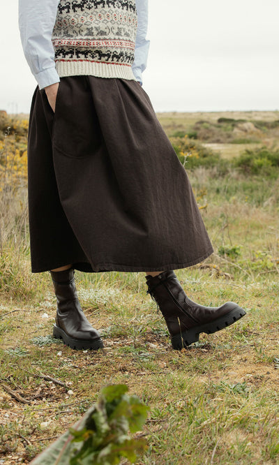 Plümo- Person in a black skirt, patterned vest, and Paola Boots standing in a grassy outdoor field.