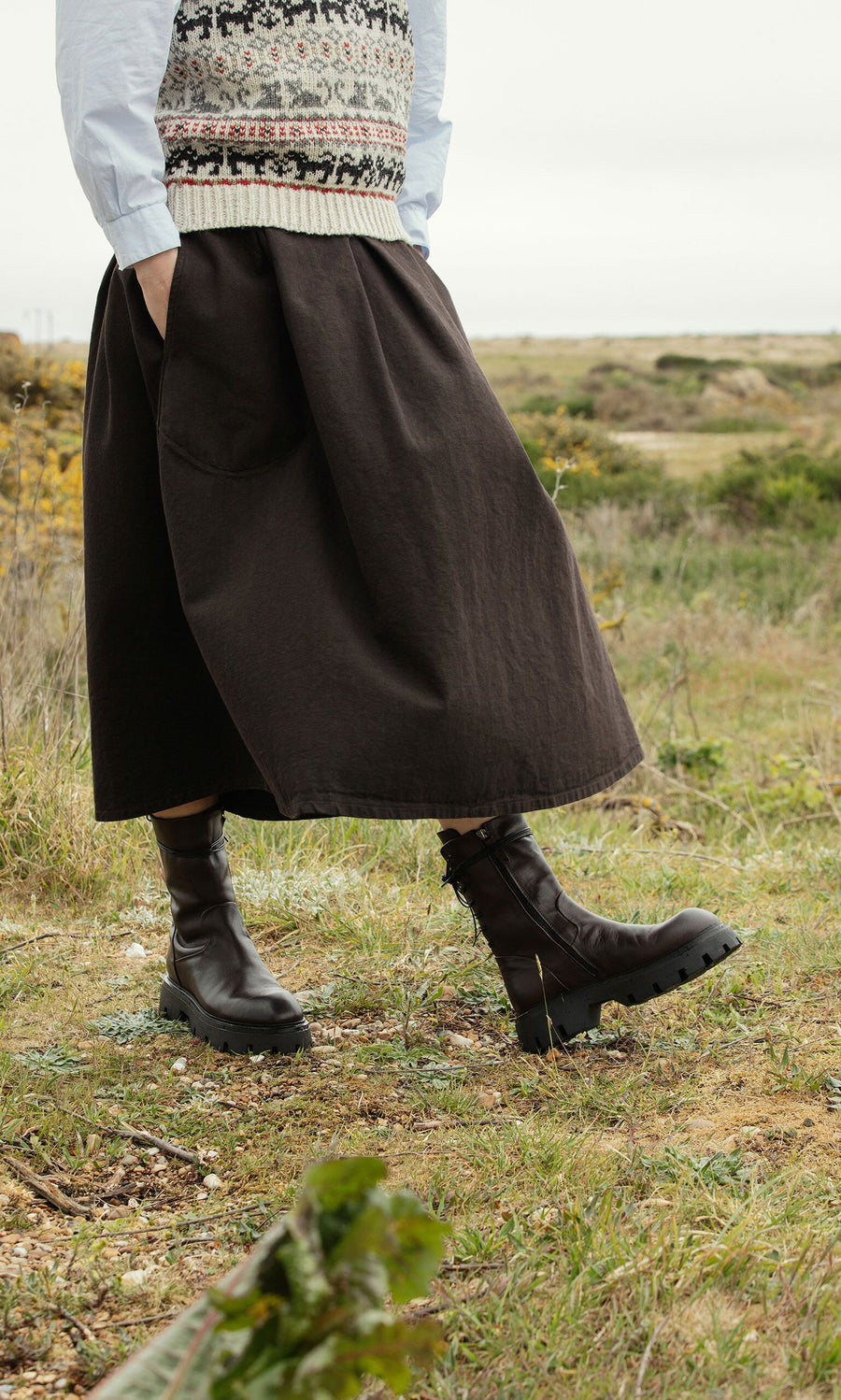 Plümo- Person in a black skirt, patterned vest, and Paola Boots standing in a grassy outdoor field.