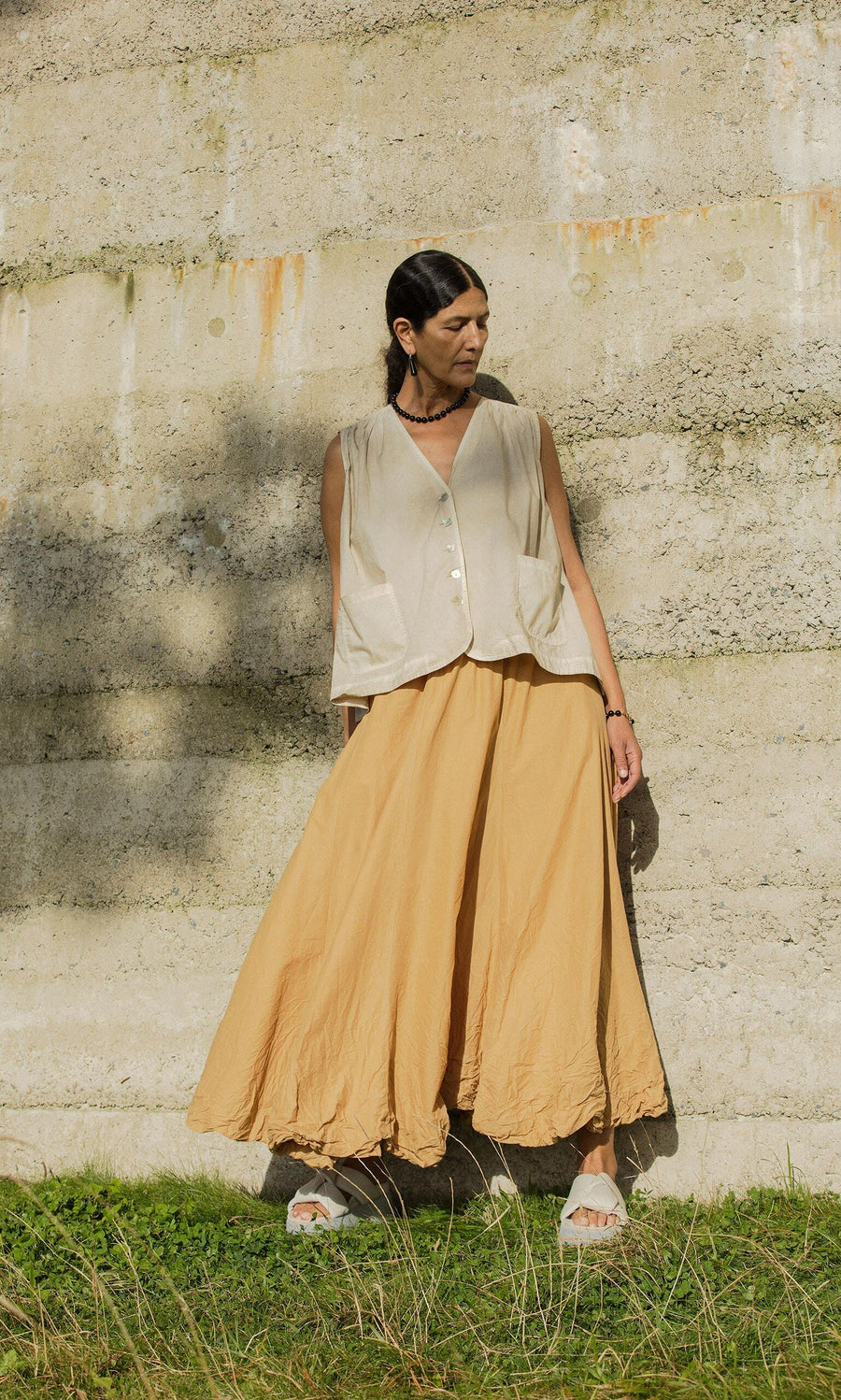 Plümo- A person stands by a concrete wall in a beige vest and the handmade Kerze Skirt, crafted in Hamburg.