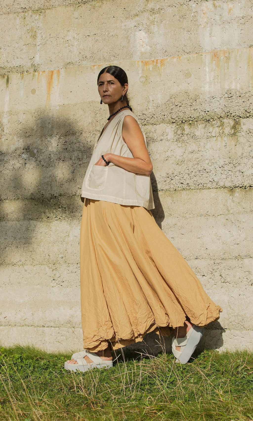 Plümo- Wearing the Cayte Waistcoat and a tan skirt, a person stands outdoors by a textured concrete wall on grassy ground.