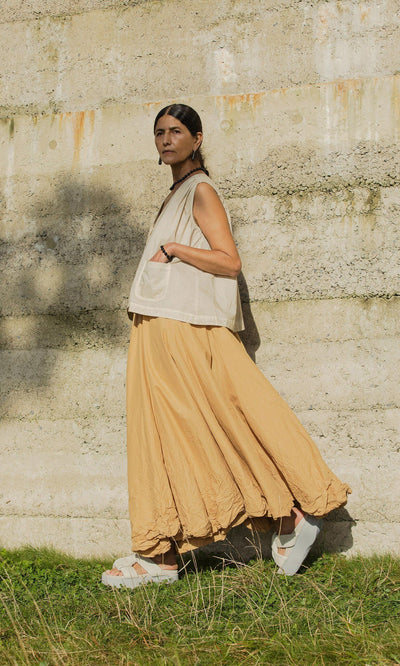 Plümo- Wearing the Cayte Waistcoat and a tan skirt, a person stands outdoors by a textured concrete wall on grassy ground.