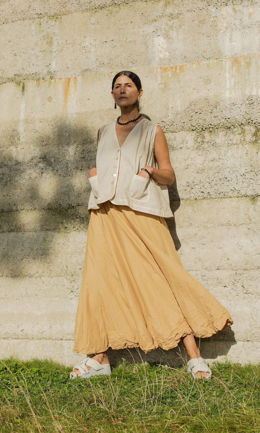 Plümo- Person in tan skirt, Cayte Waistcoat with shell buttons, and sandals stands on grass by a textured concrete wall confidently.