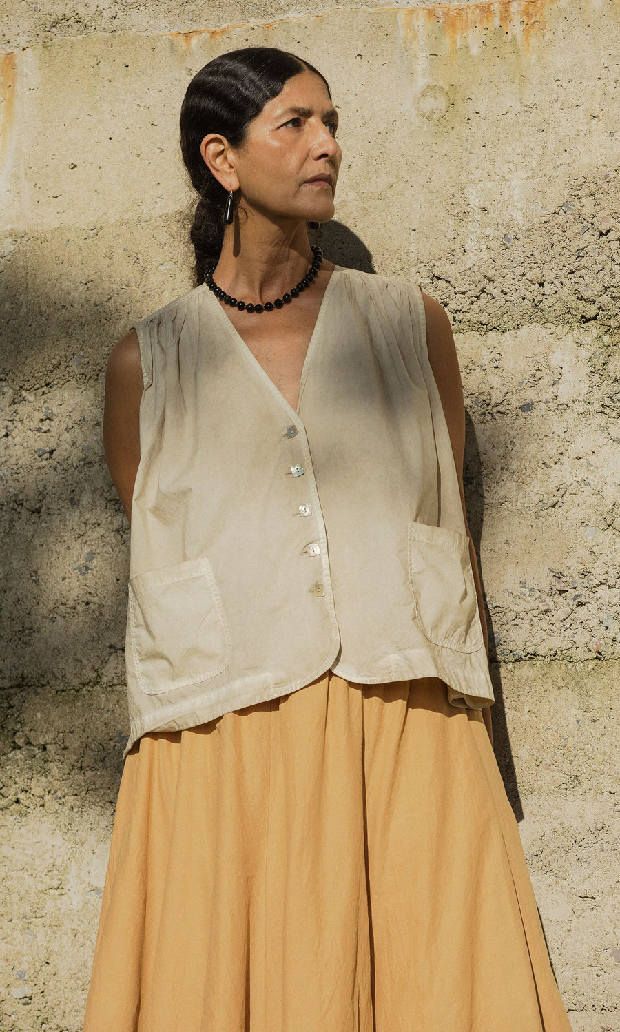 Plümo- Woman in the Cayte Waistcoat and tan skirt stands against a textured concrete wall, looking to the side.