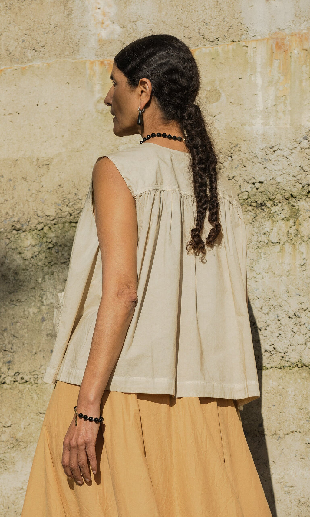 Plümo- Woman with braided hair wears the Cayte Waistcoat, sleeveless and beige, standing by a textured concrete wall outdoors.