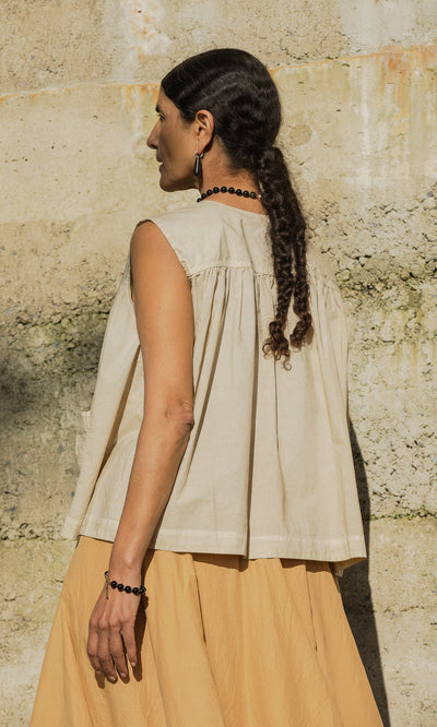 Plümo- Woman with braided hair wears the Cayte Waistcoat, sleeveless and beige, standing by a textured concrete wall outdoors.
