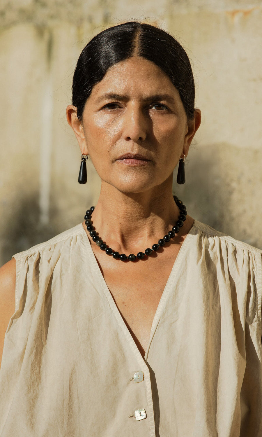 Plümo- A woman with dark hair in a bun, wearing Hera Earrings and a beige top, stands outdoors.