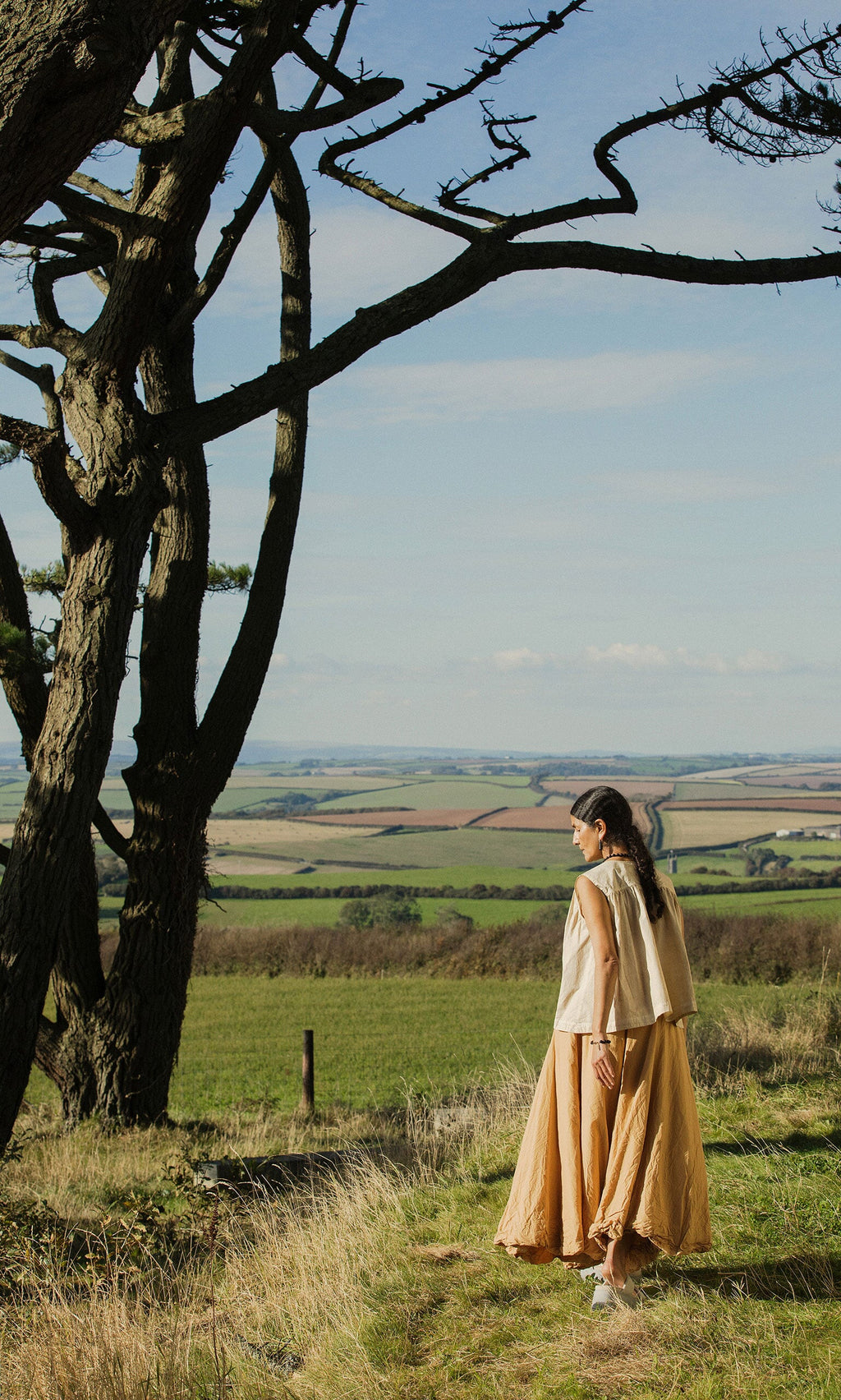 Plümo- A woman in the Cayte Waistcoat stands by a tree, looking out over green fields and distant hills.