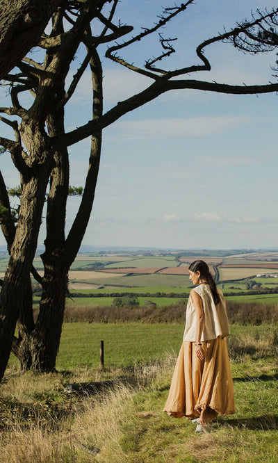 Plümo- A woman in the Cayte Waistcoat stands by a tree, looking out over green fields and distant hills.