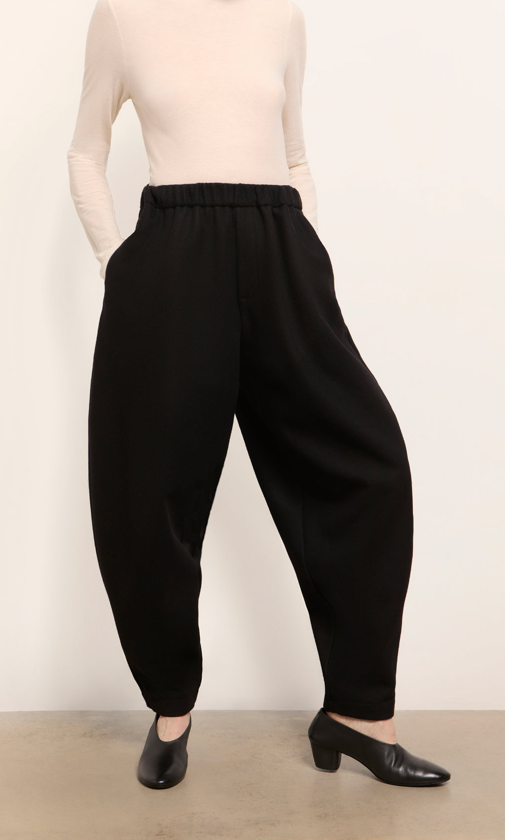 Plümo  Person wearing a cream long-sleeve top, Circle Pants - Boiled Black, and black low-heeled shoes on a gray floor.