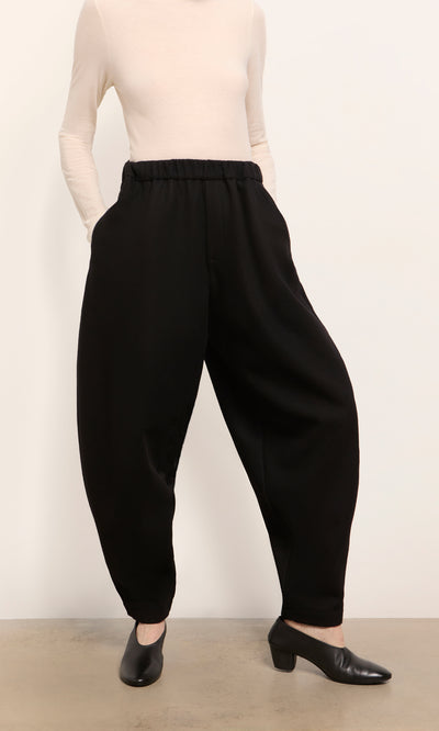 Plümo  Person wearing a cream long-sleeve top, Circle Pants - Boiled Black, and black low-heeled shoes on a gray floor.