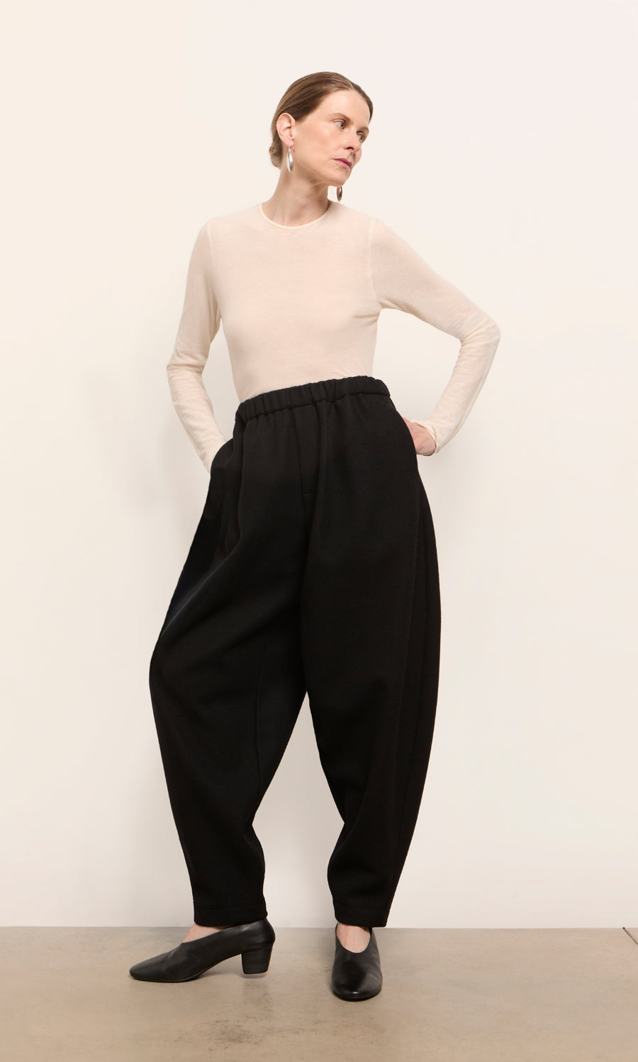 Plümo  Woman in a beige top and Circle Pants - Boiled Black stands with hands in pockets, gazing off to the side.