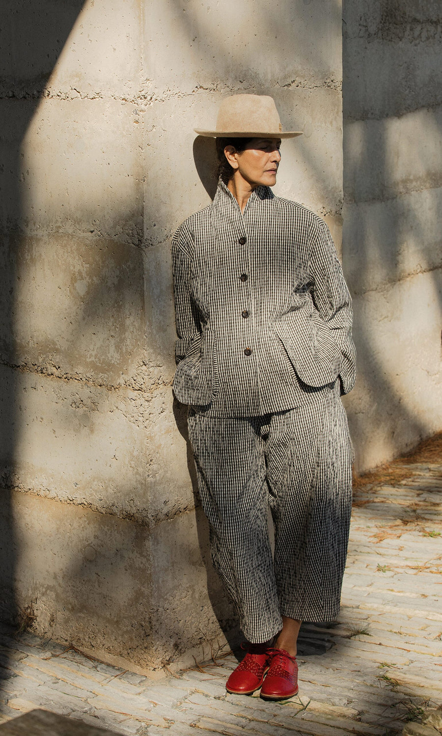 Plümo- Person in Butoni Pants - Gingham and a tan hat leans on a wall, bright red shoes catching sunlight.