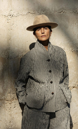 Plümo- Person in the Gardeners Jacket - Gingham and a tan hat stands against a stone wall, hands in pockets, facing the camera.