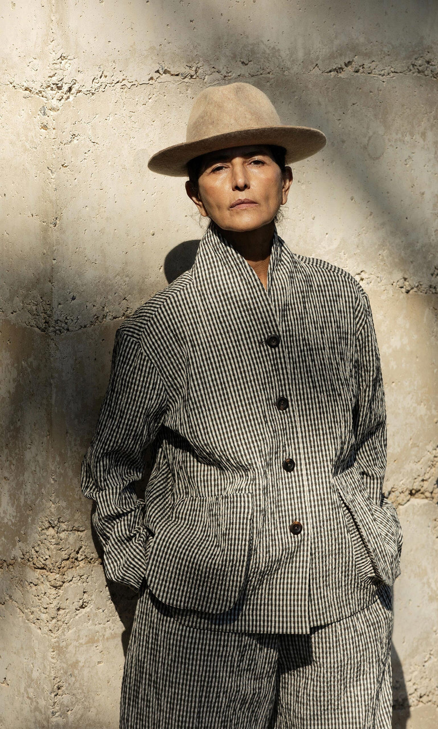 Plümo- Person in the Gardeners Jacket - Gingham and a tan hat stands against a stone wall, hands in pockets, facing the camera.