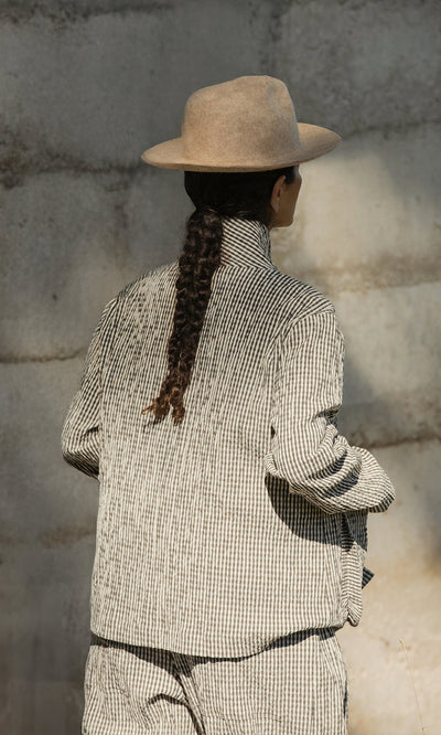 Plümo  Person with long braid in a checked outfit wears the Noma Felted Hat, facing a textured stone wall.
