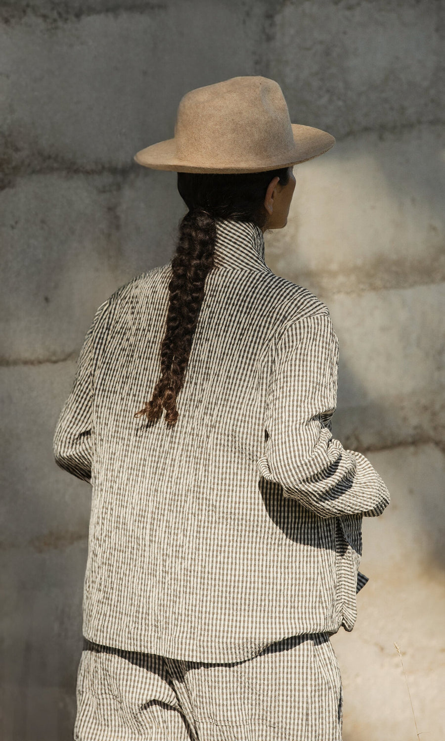Plümo- Person with a long braid in a Gardeners Jacket - Gingham and tan hat stands facing a sunlit concrete wall.