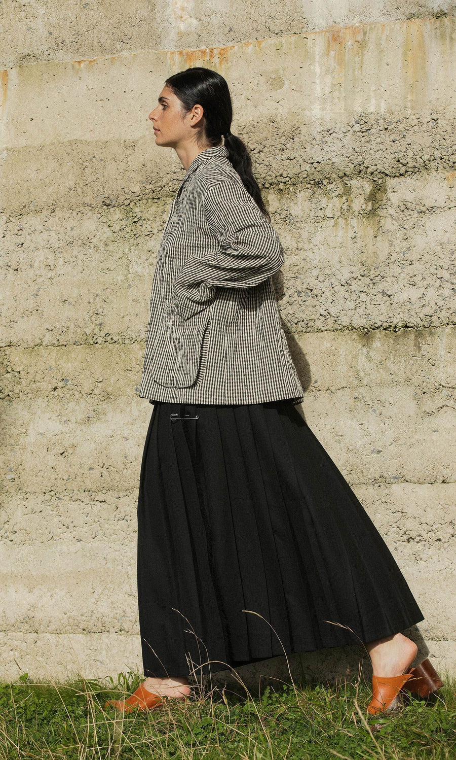 Plümo- Person in a Gardeners Jacket - Gingham, black pleated skirt, and brown shoes walks past a textured concrete wall on grass.