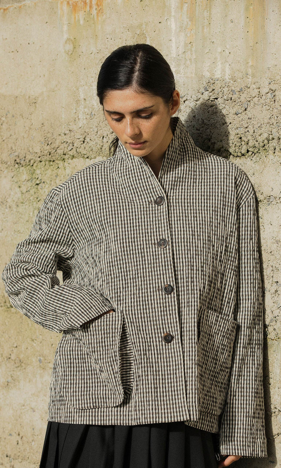 Plümo- Woman in a Gardeners Jacket - Gingham, made in London, stands by a sunlit concrete wall, looking downward.