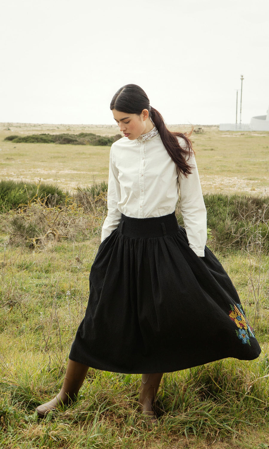 Plümo- Woman wears the Cleo Shirt with a black skirt, standing in a grassy field with hands in her skirt pockets.