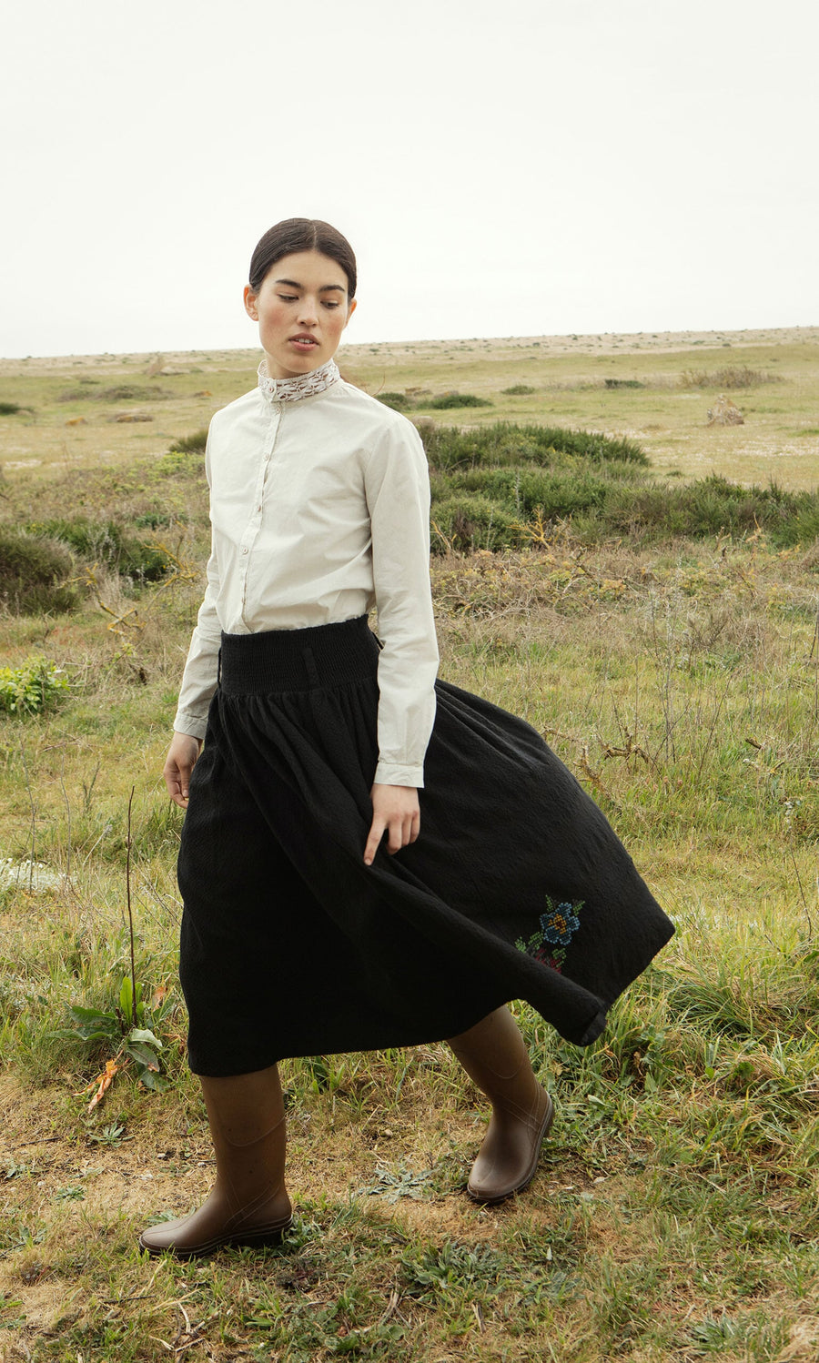 Plümo- A woman in a Cleo Shirt and black skirt stands in a grassy field, looking to the side, wearing brown boots.