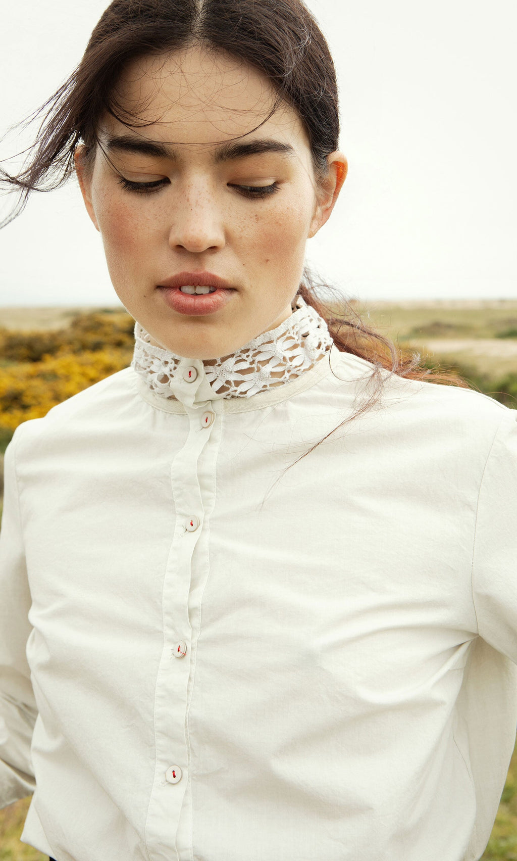 Plümo- Woman with dark hair wears the Cleo Shirt with lace collar outdoors, looking down with a neutral expression.