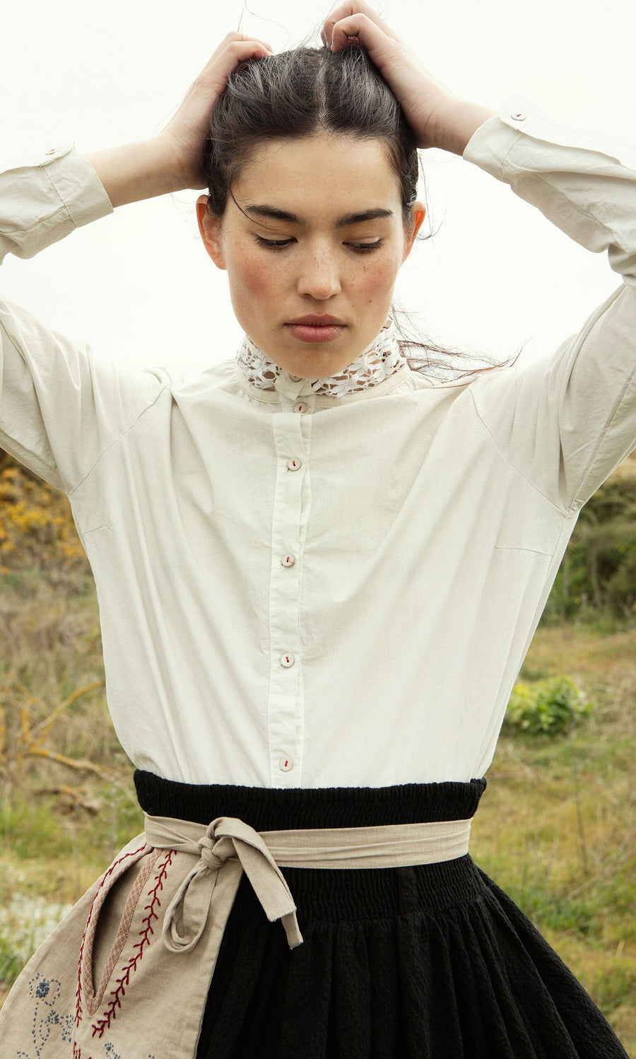 Plümo- A person in a Cleo Shirt and black skirt stands outdoors with hands behind head and eyes closed.