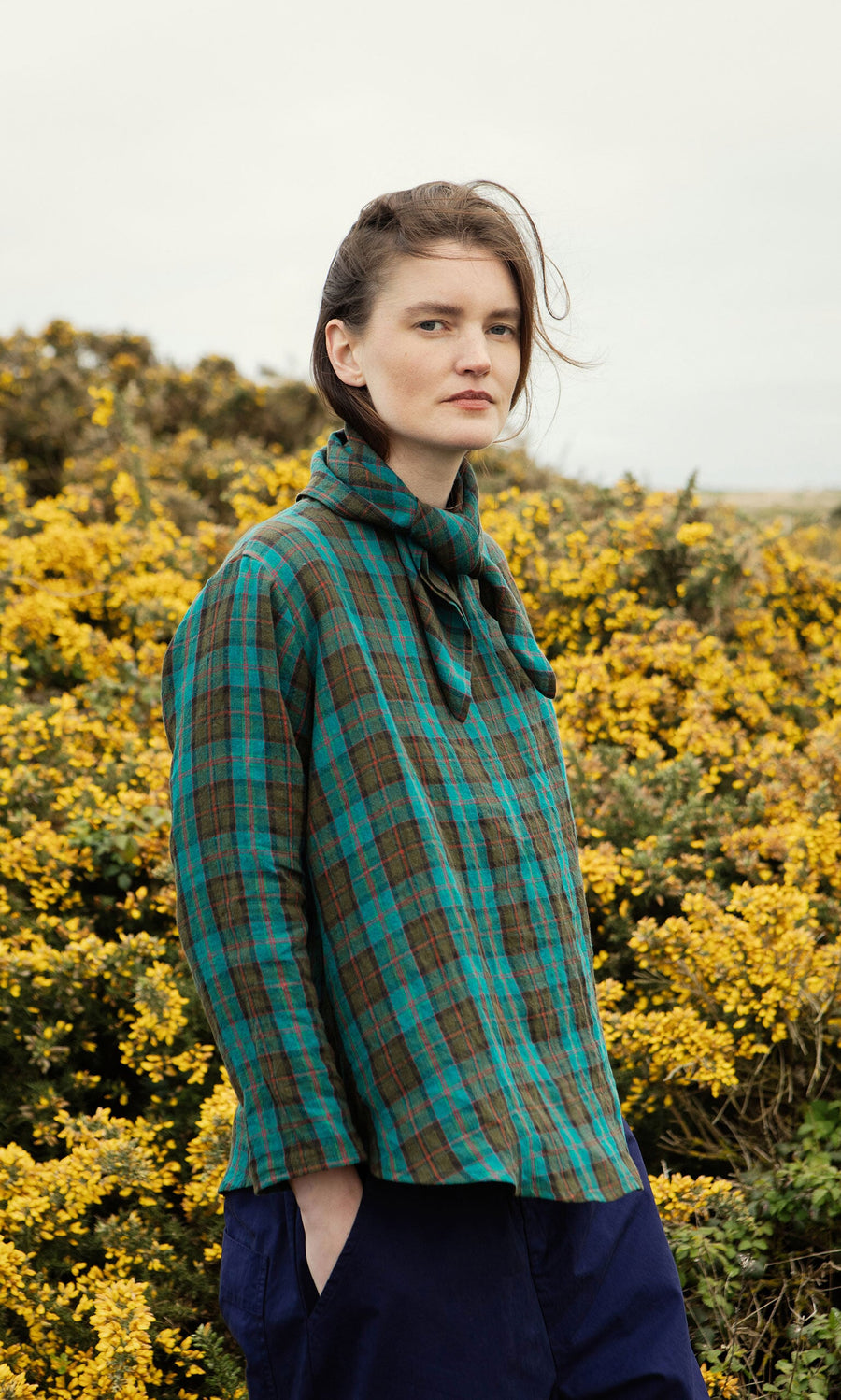 Plümo- Someone wears the Clodagh Blouse in green tartan, standing before yellow flowering bushes on a cloudy day.