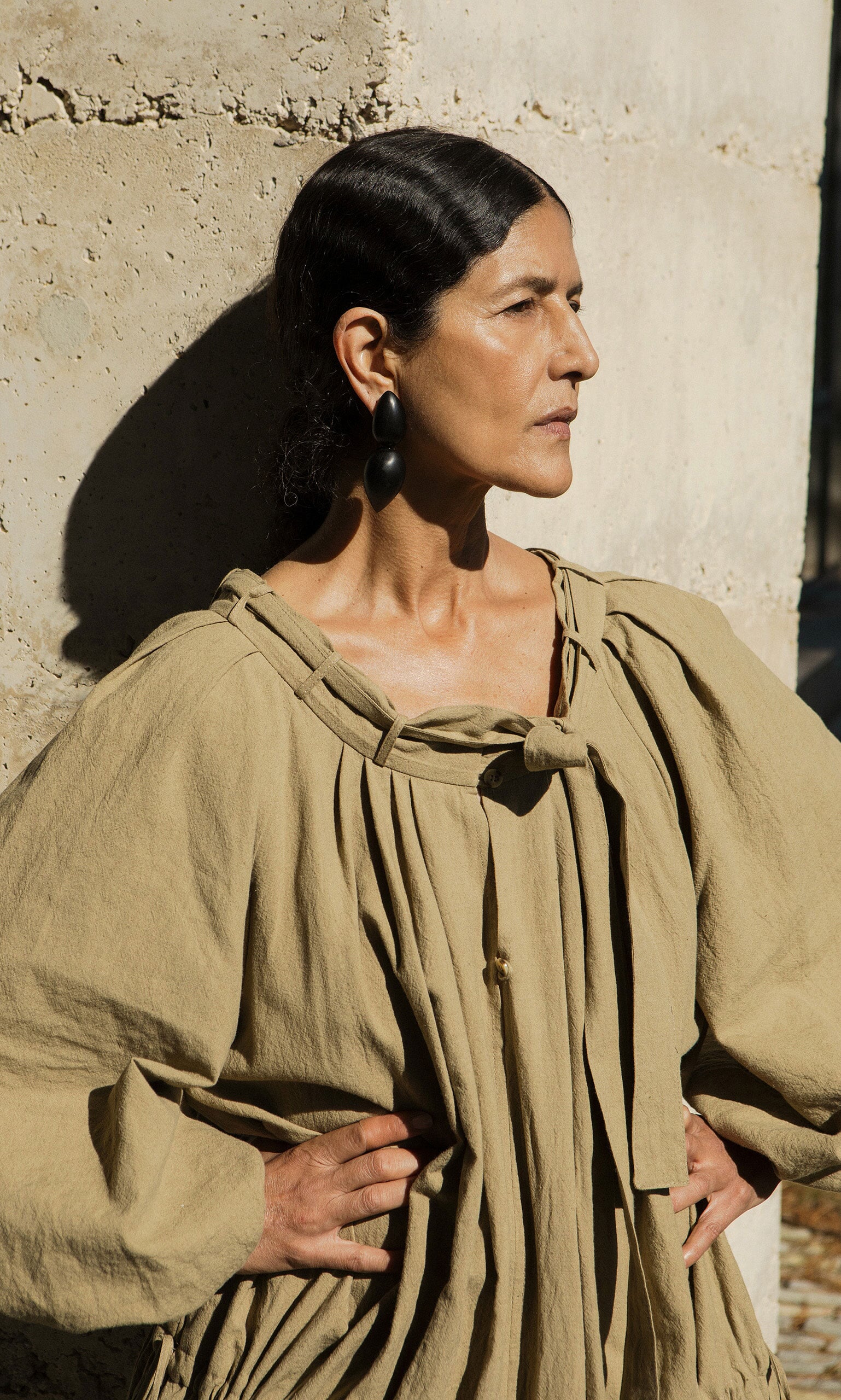 Plümo- Woman in a tan oversized dress stands against a textured wall, showcasing Coalvale Earrings with hands on her hips.