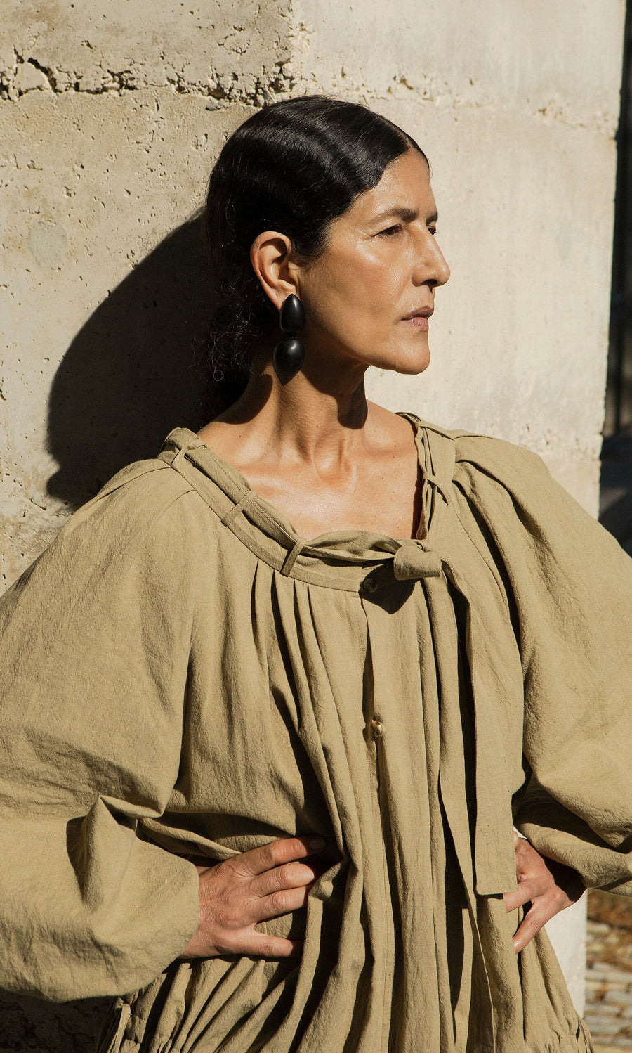 Plümo- Woman in a tan oversized dress stands against a textured wall, showcasing Coalvale Earrings with hands on her hips.