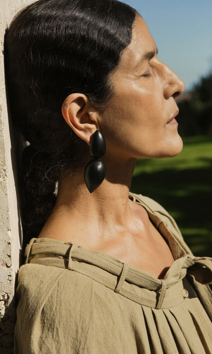 Plümo- Woman in a beige top leans against a wall, eyes closed, sunlight on her face, wearing Coalvale Earrings.