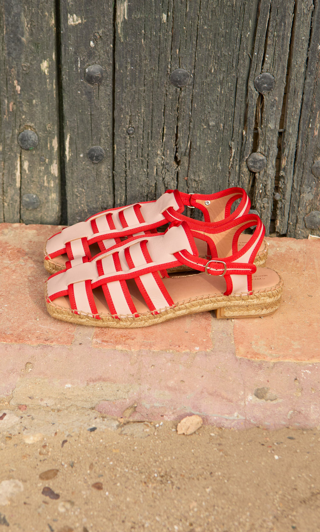 Plümo- Cordoba Espadrilles—red leather sandals with jute soles—rest against a rustic wooden door on tiles.