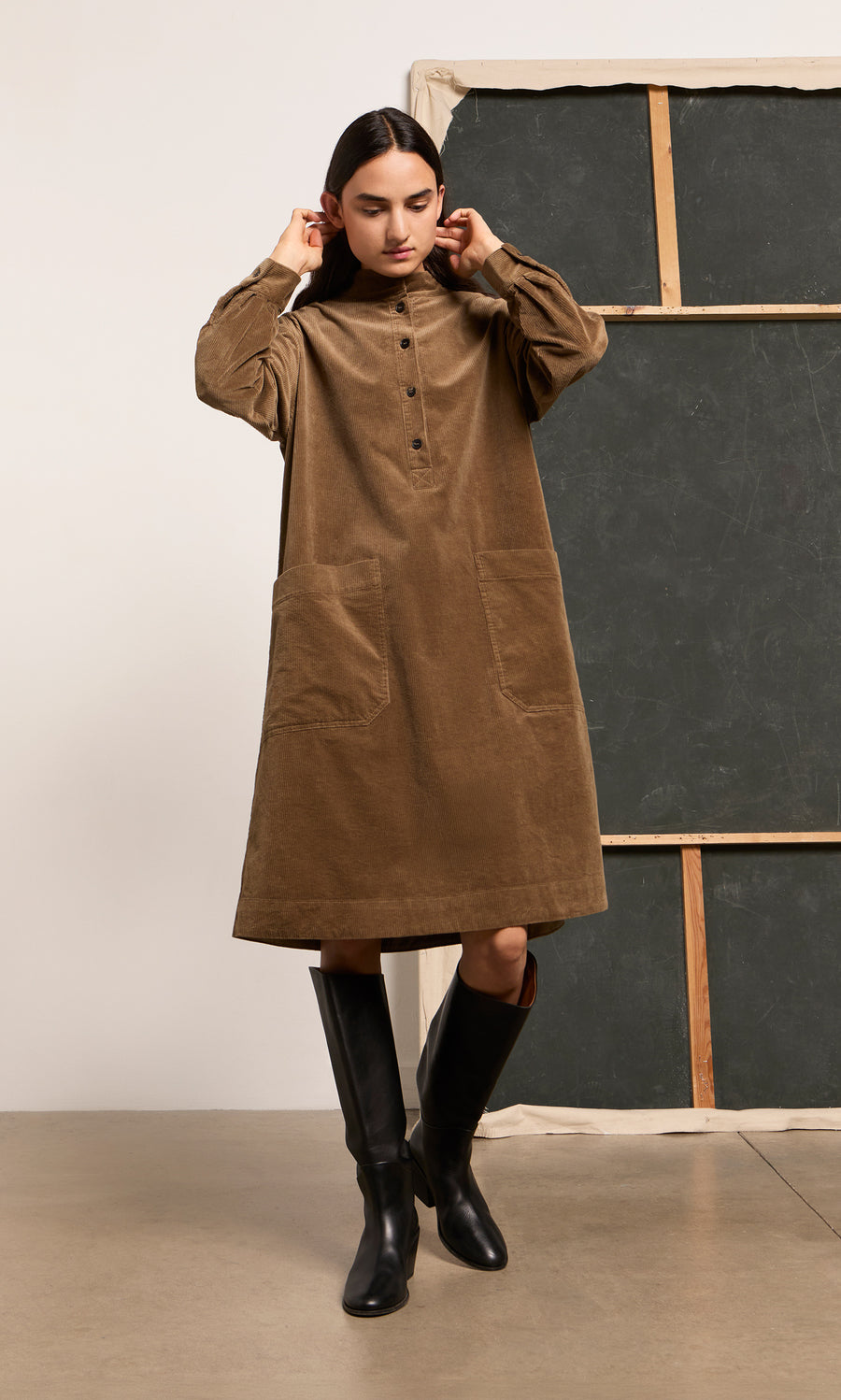 Plümo  Woman in a midi-length Frances Dress with large pockets and black boots stands indoors near a chalkboard.