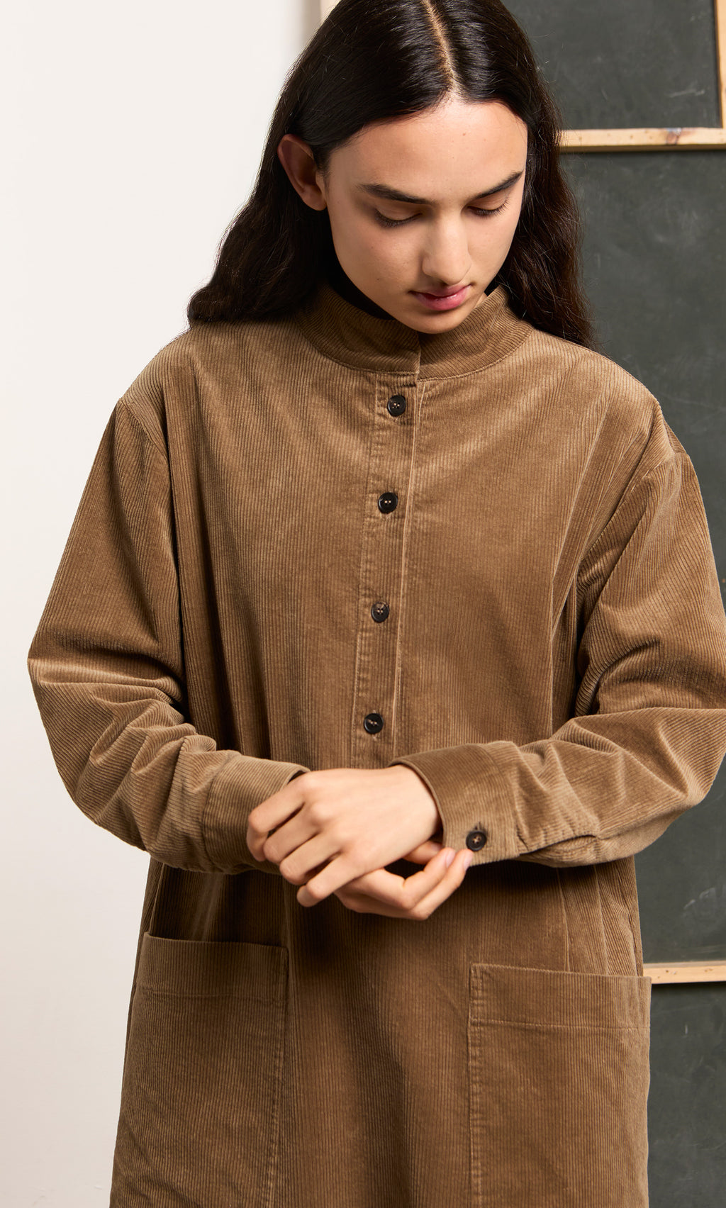Plümo  A person with long dark hair wears the Frances Dress—a brown, midi cotton corduroy dress with two large front pockets.