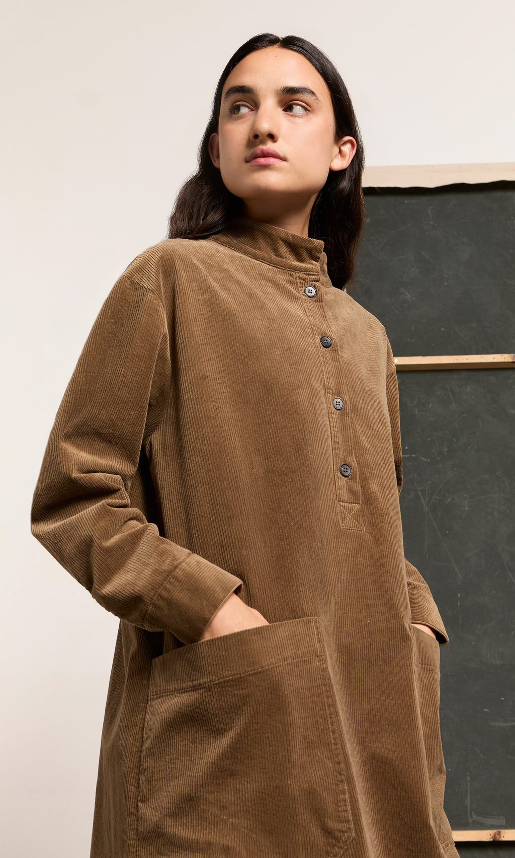 Plümo  Person wearing the Frances Dress, a brown cotton corduroy style with pockets, standing by a blackboard, looking aside.