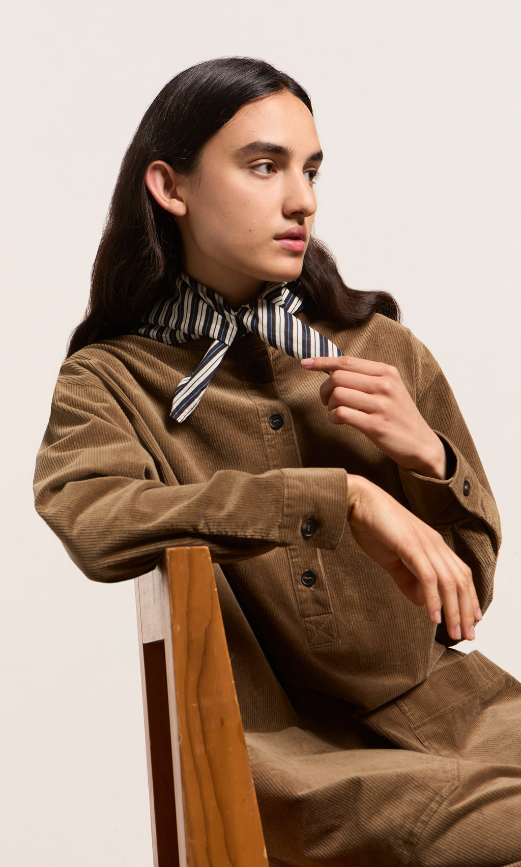 Plümo  Person in brown corduroy sits sideways, wearing a Paris atelier-inspired blue stripe necktie and looking right.
