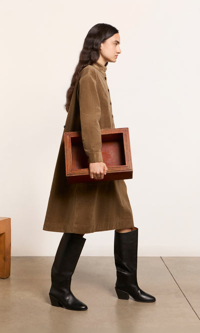 Plümo  Woman in a Frances Dress and black boots walks, holding a large box against a plain background.