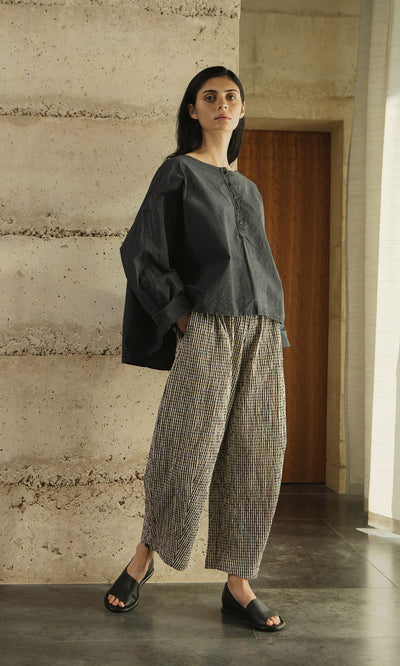 Plümo- Woman in loose dark top, Butoni Pants - Gingham, and black sandals stands indoors by a textured wall and wooden door.