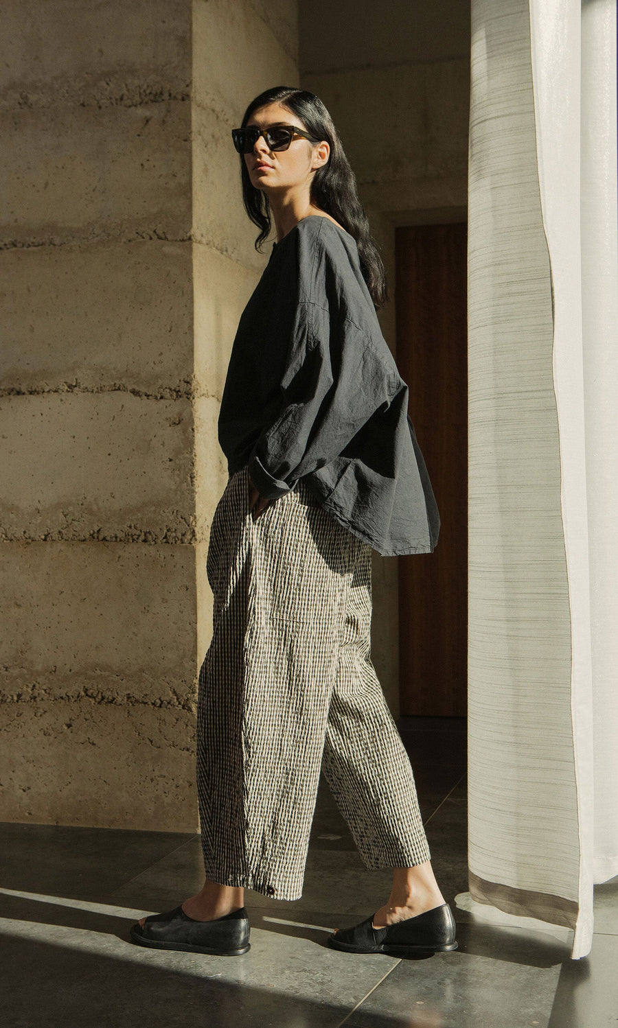 Plümo- Person in sunglasses wears the Corie Blouse with a boxy fit and checked pants, standing in sunlight by a curtain.
