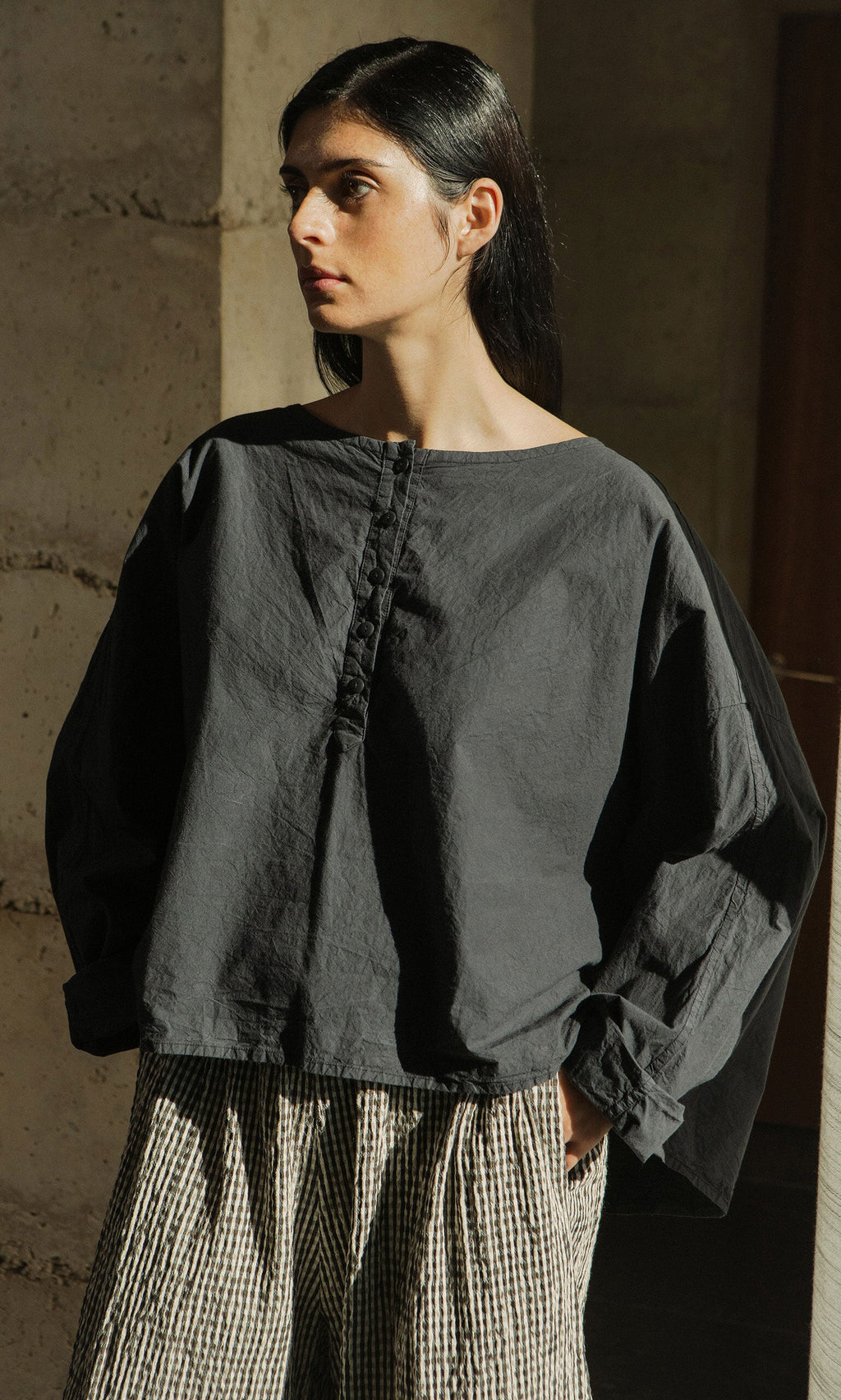 Plümo- Someone in a boxy silhouette Corie Blouse and checkered pants stands in sunlight, looking left with hands in pockets.