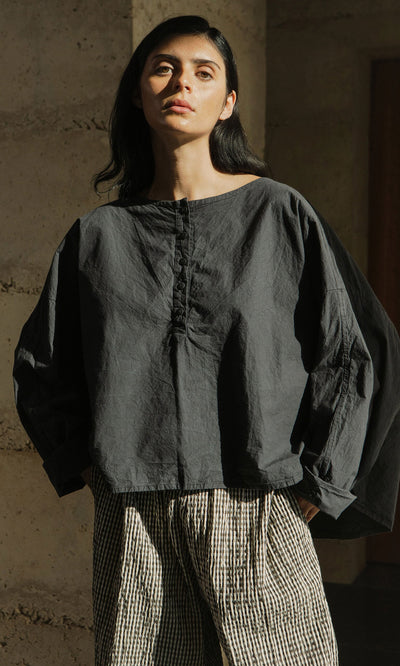 Plümo- Woman in a boxy silhouette Corie Blouse stands in sunlight against a textured, neutral wall wearing checkered pants.
