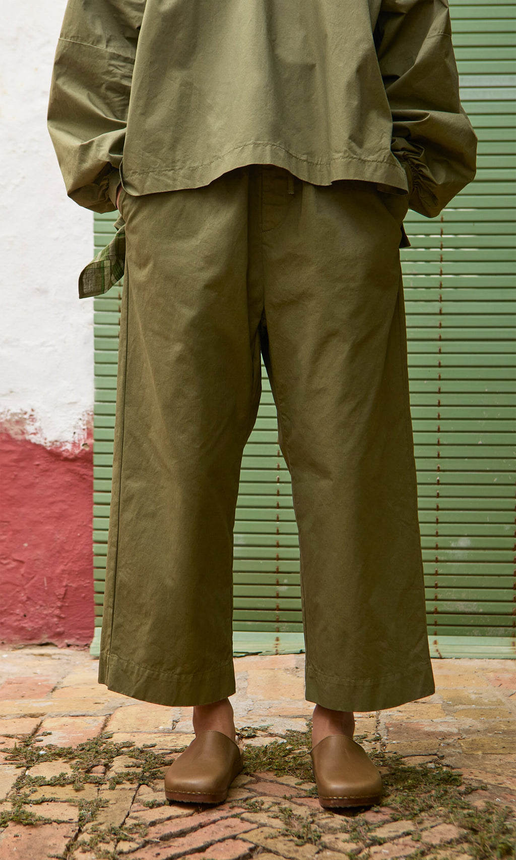 Plümo- Person wearing Calke Green Pants, matching top, and brown clogs on a patterned tile floor.