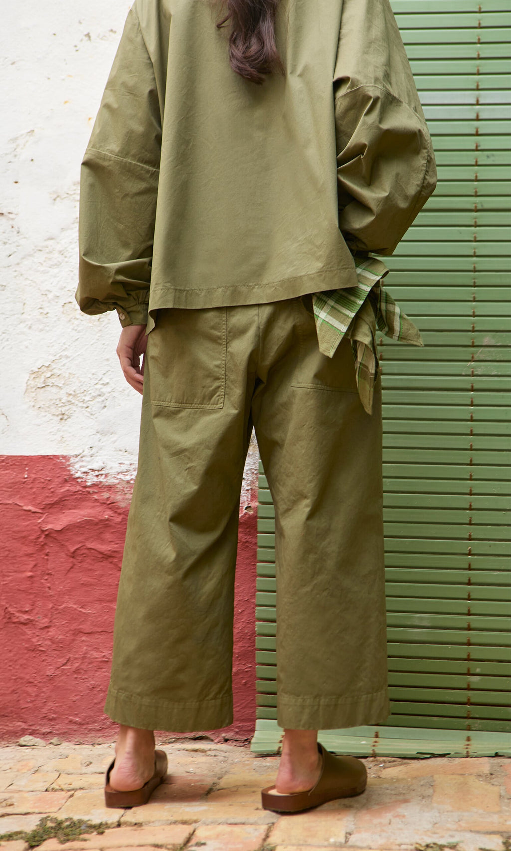 Plümo- Person wearing Calke Green Pants and sandals, standing on a brick floor with plaid cloth in pocket.