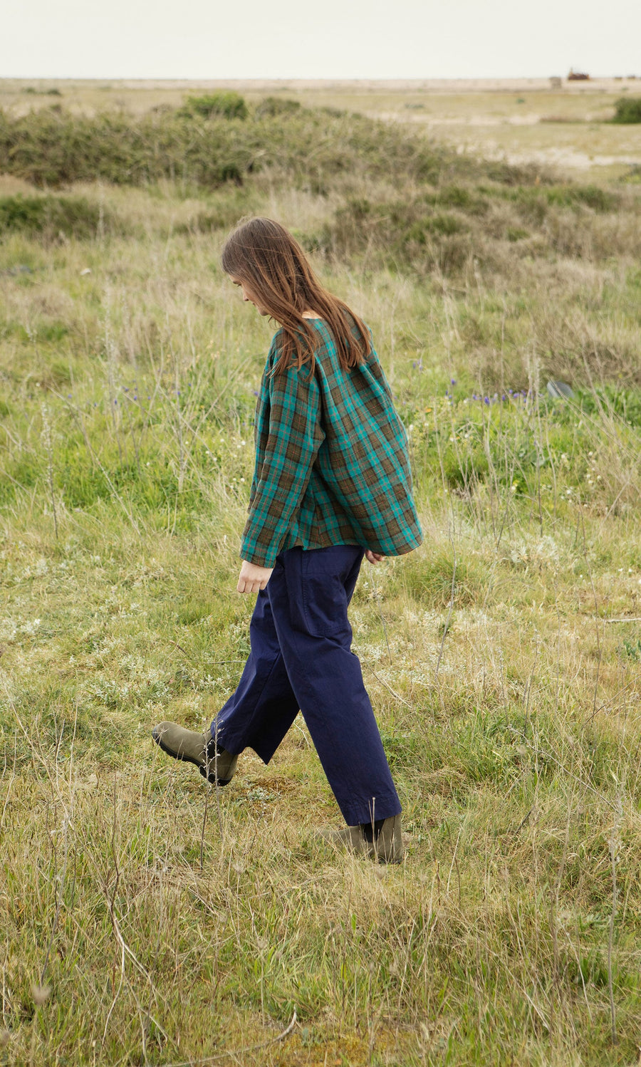 Plümo- Someone in a green Clodagh Blouse and blue pants walks through tall grass in a field.