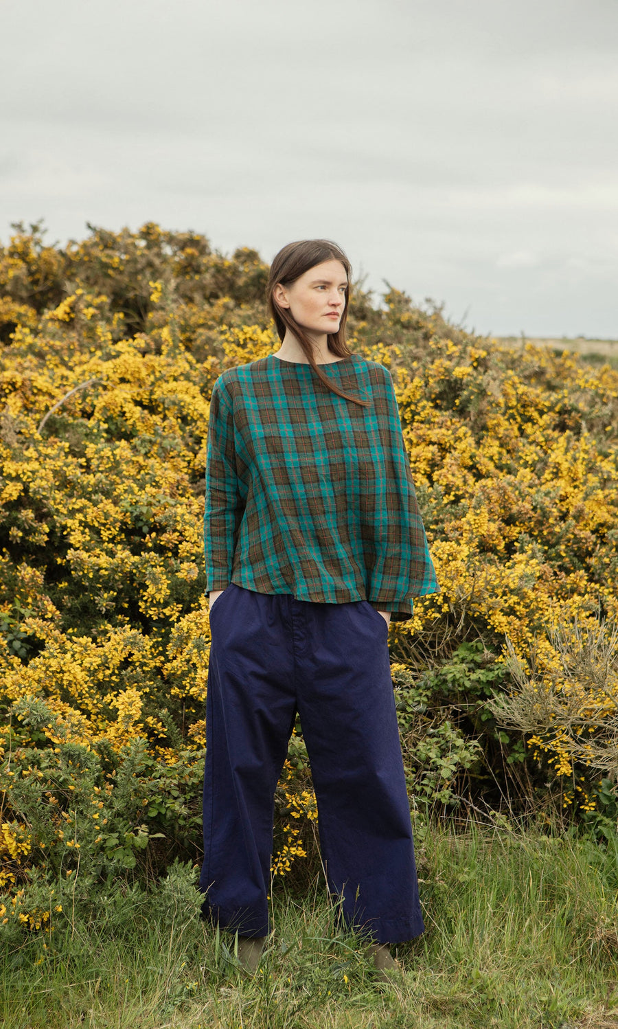 Plümo- Woman wears the Clodagh Blouse and wide navy pants while standing by yellow-flowering bushes outdoors.