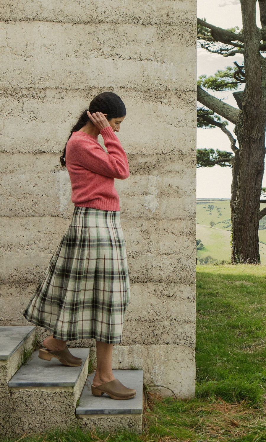 Plümo- Woman in pink sweater and Hawksmoor Kilt stands on outdoor steps in Dublin beside a concrete wall.