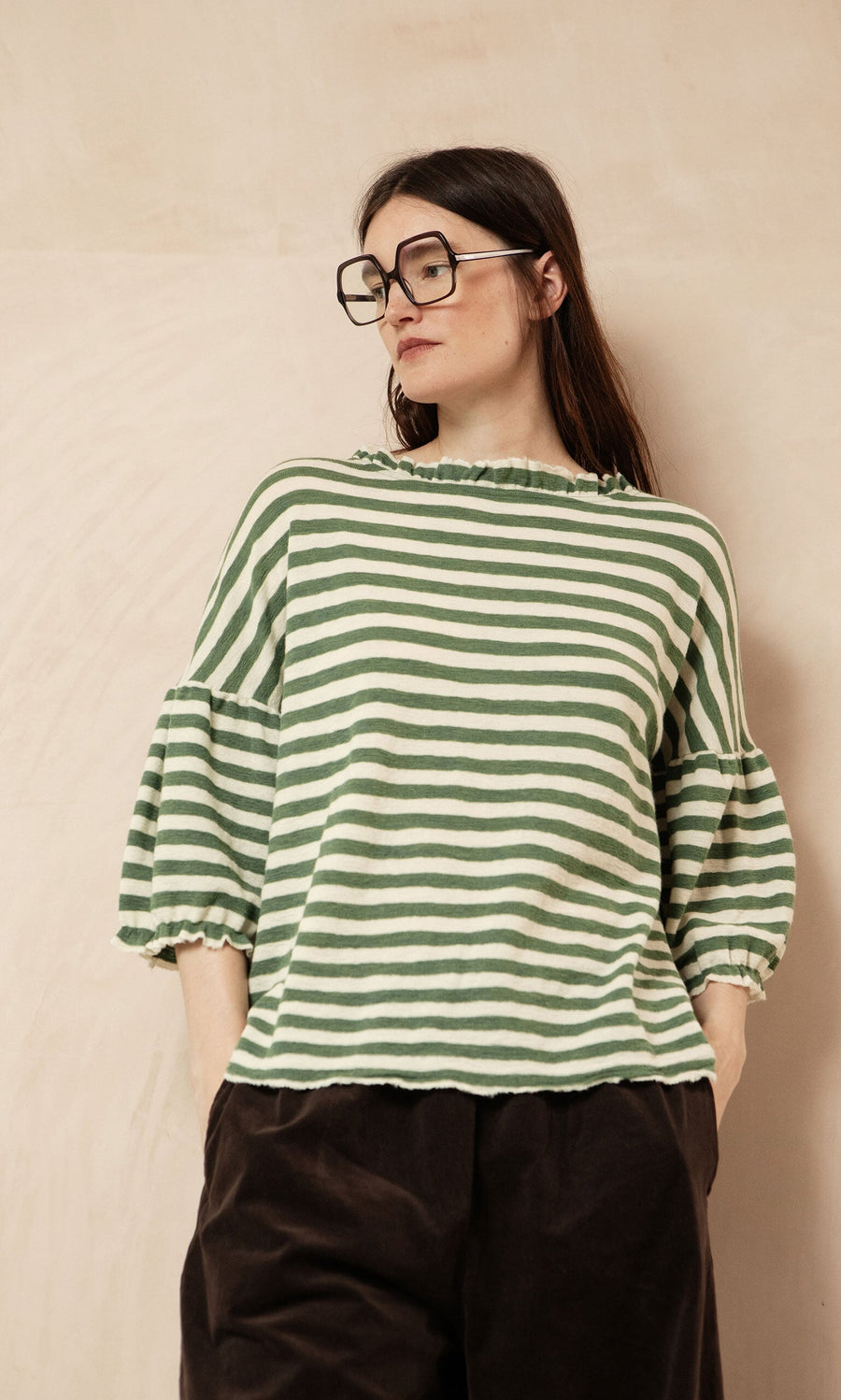 Plümo- Woman wears Square Readers and a green striped ruffle-sleeve top, standing against a beige wall.
