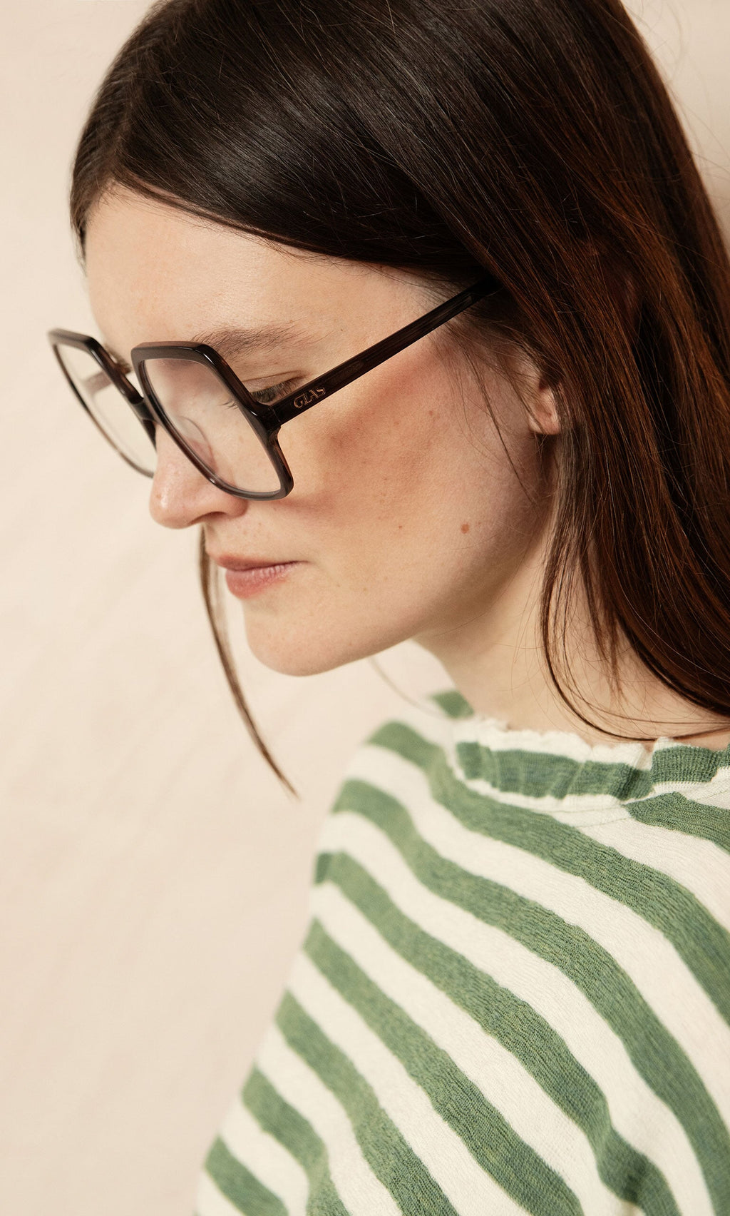Plümo- Woman with long brown hair wears Square Readers and a green-and-white striped sweater, looking downward.