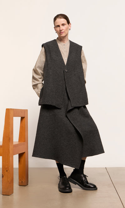 Plümo  Person in an Eardley Skirt and vest over a beige shirt, standing by a wooden chair on a light floor and white wall.