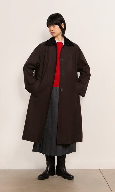 Plümo  Person in a long brown Doherty Coat - Soil, red sweater, gray skirt, and black boots stands against a plain wall.