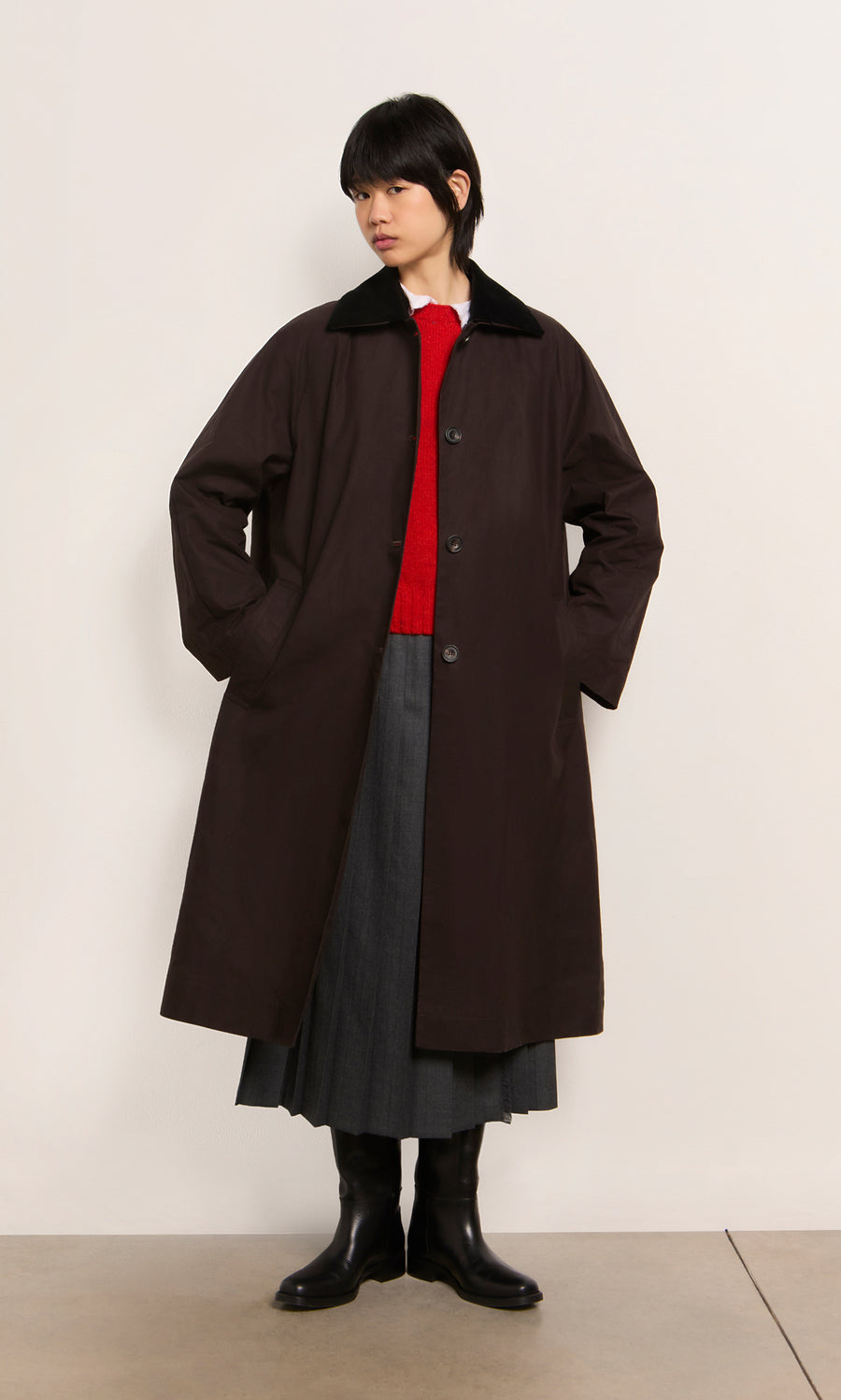 Plümo  Person in a long brown Doherty Coat - Soil, red sweater, gray skirt, and black boots stands against a plain wall.
